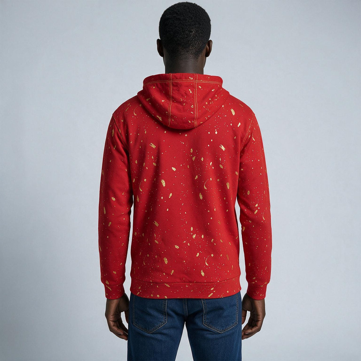 A man viewed from behind wearing a red pullover hoodie with a gold speckled pattern, paired with blue jeans, against a light gray studio background.