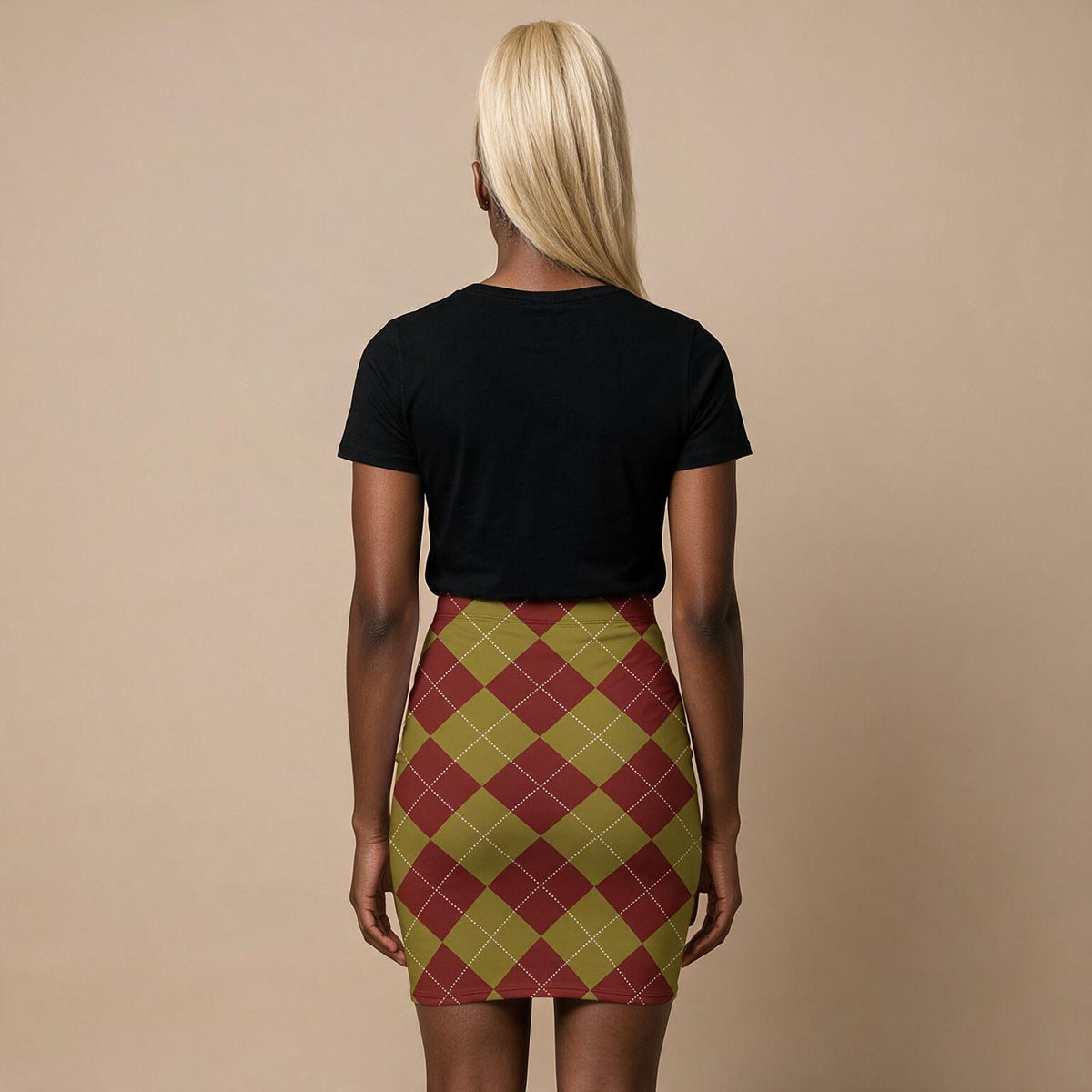 A woman viewed from behind wearing a black short-sleeve t-shirt and a fitted mini skirt with an argyle pattern in olive green, burgundy, and cream. She is standing against a solid beige studio background.