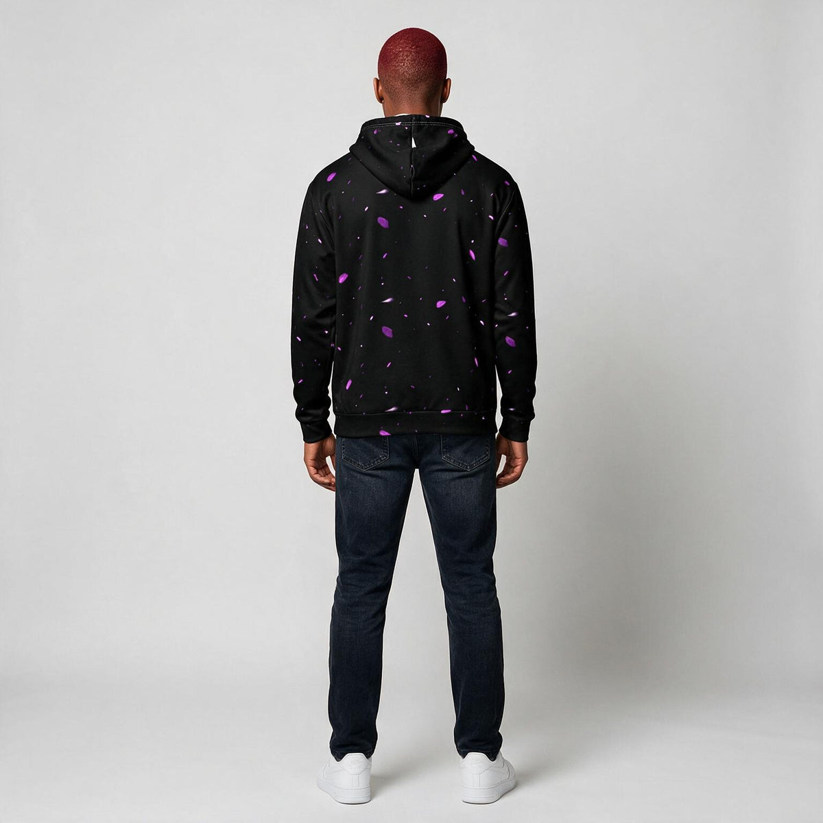 A man viewed from behind wearing a black pullover hoodie with a purple and white confetti pattern, paired with dark blue jeans and white sneakers, against a light gray studio background.