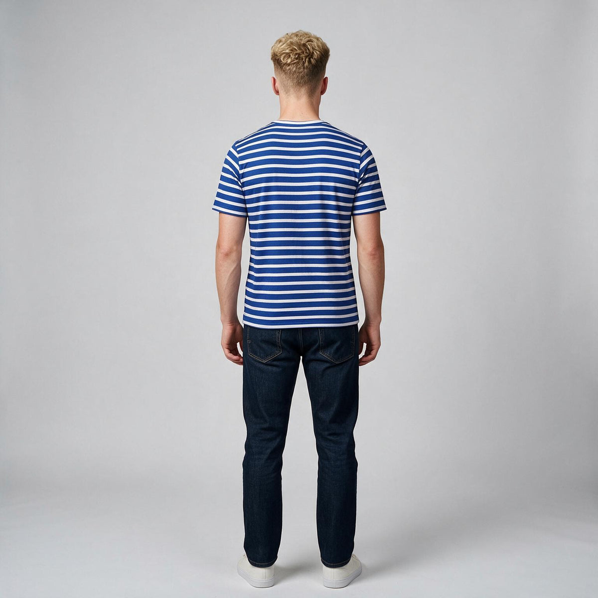 A man viewed from behind wearing a blue and white horizontally striped short-sleeve t-shirt, dark blue jeans, and white sneakers, standing against a light gray studio background.
