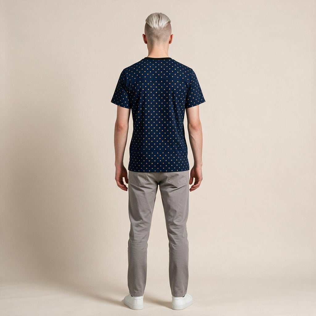 A man viewed from behind wearing a navy blue short-sleeve t-shirt with a small gold geometric pattern, paired with light gray trousers and white sneakers, against a solid beige studio background.