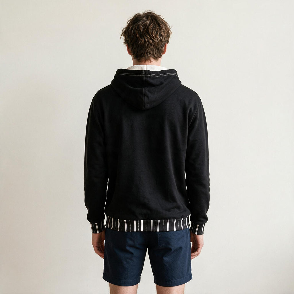 A man viewed from behind wearing a black pullover hoodie with striped ribbed trim at the hood, cuffs, and hem, paired with navy shorts against a plain off-white background.