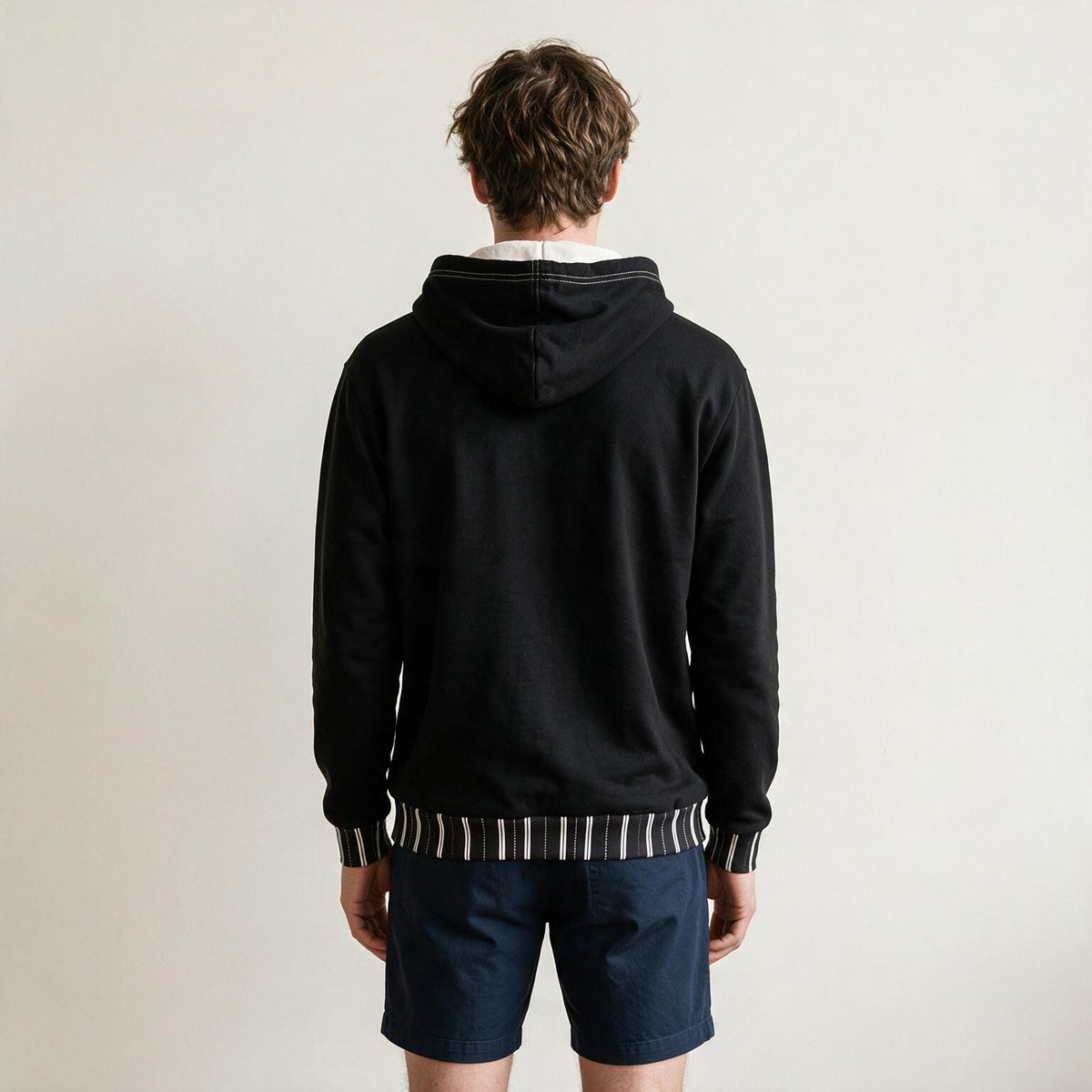 A man viewed from behind wearing a black pullover hoodie with striped ribbed trim at the hood, cuffs, and hem, paired with navy shorts against a plain off-white background.