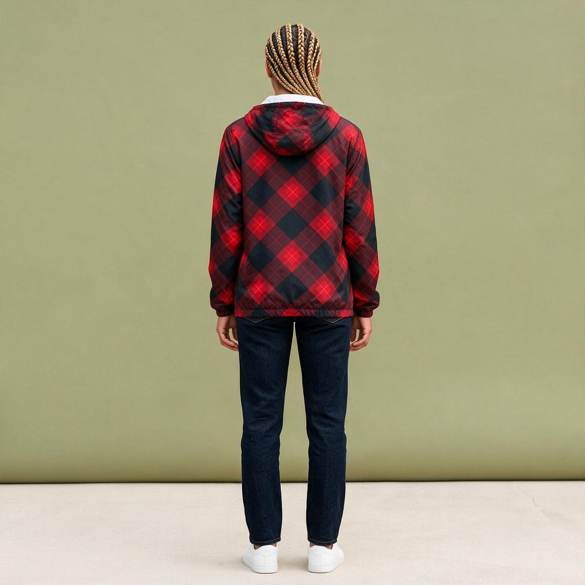 A person viewed from the back wearing a red and black buffalo plaid hooded jacket, dark blue jeans, and white sneakers, standing against a solid sage green wall.