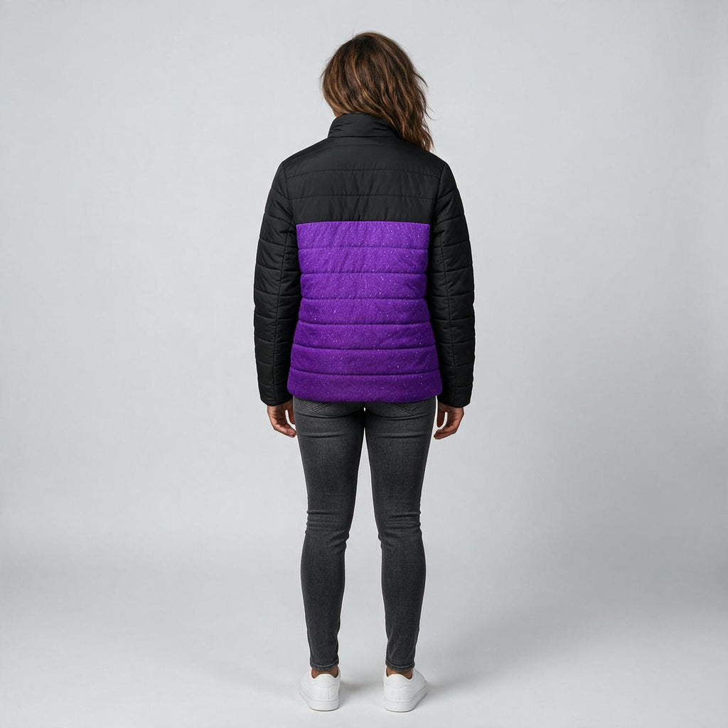 A woman viewed from the back wearing a black puffer jacket with a vibrant purple horizontal quilted panel across the middle, paired with dark gray skinny jeans and white sneakers, against a light gray studio background.