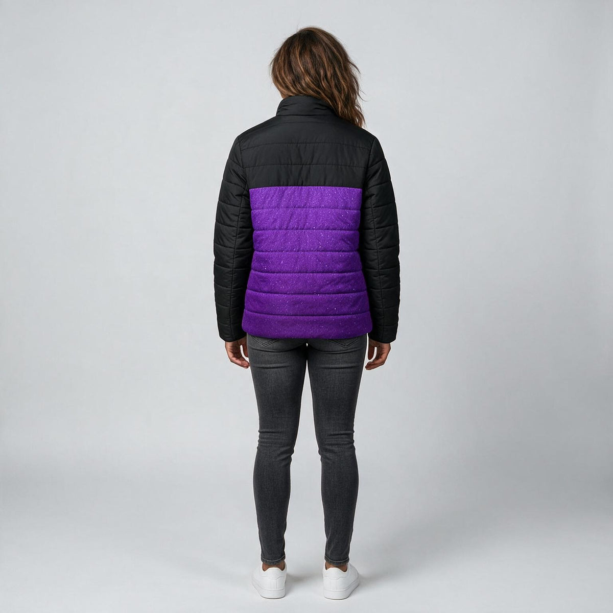 A woman viewed from the back wearing a black puffer jacket with a vibrant purple horizontal quilted panel across the middle, paired with dark gray skinny jeans and white sneakers, against a light gray studio background.