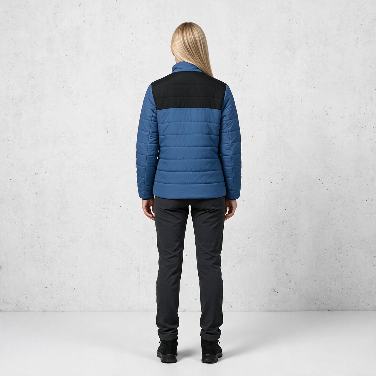 A woman viewed from behind wearing a blue and black puffer jacket, dark gray pants, and black boots, standing against a light gray textured studio wall.