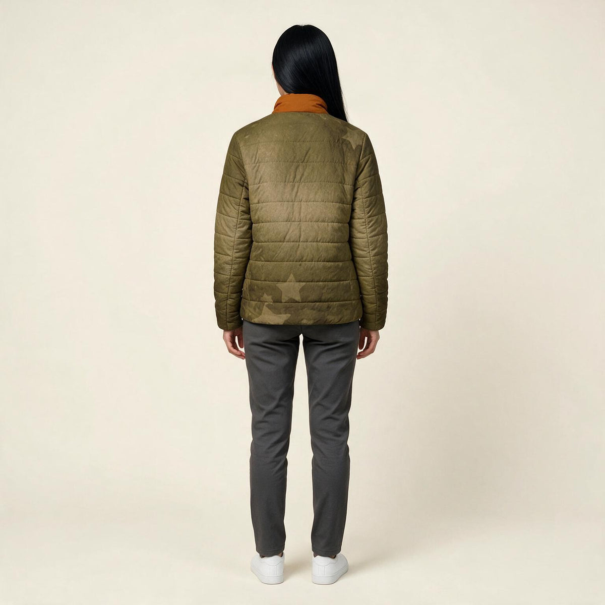 A woman viewed from behind wearing an olive green quilted puffer jacket with a contrasting orange collar, dark gray pants, and white sneakers, set against a plain beige studio background.