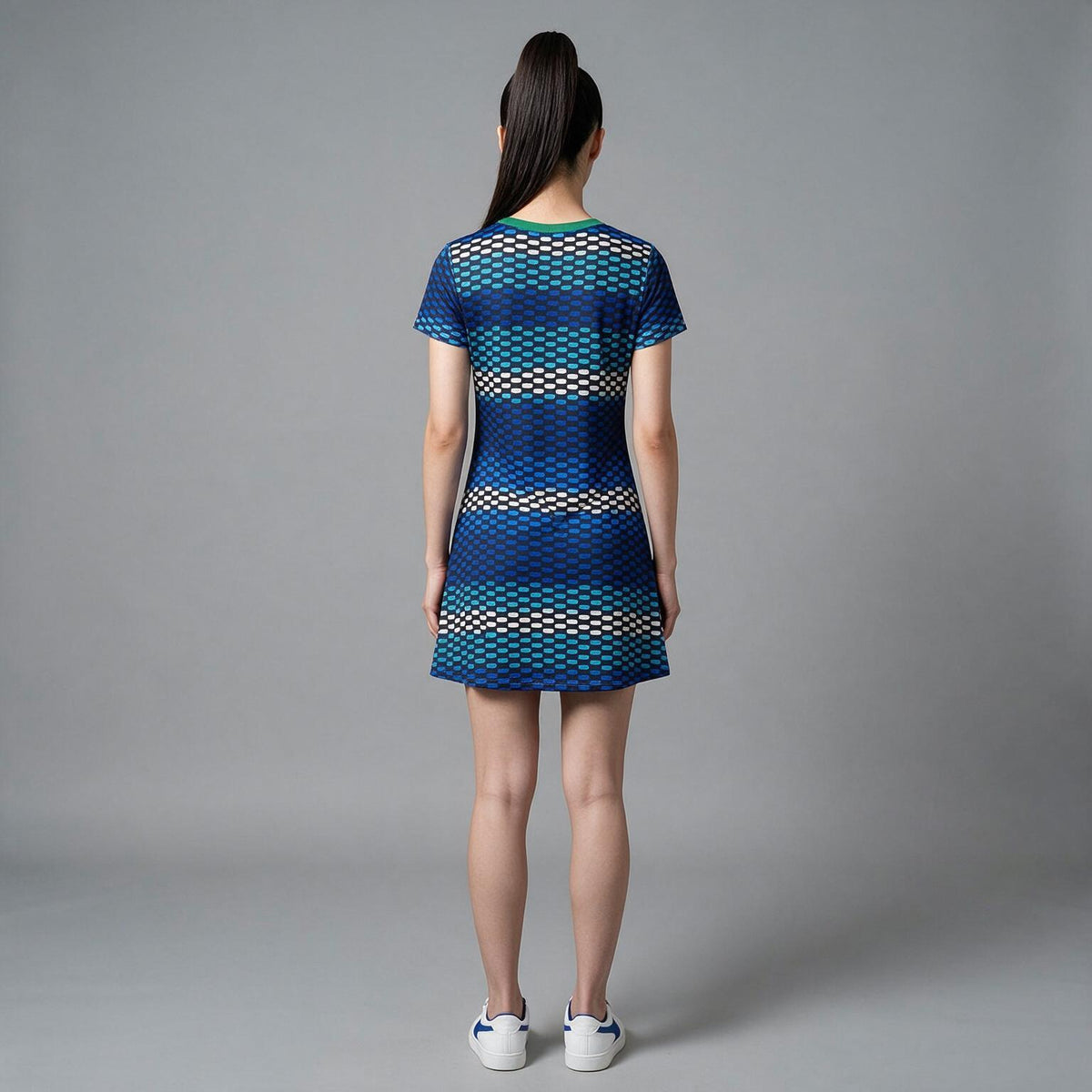 A woman viewed from behind wearing a short-sleeve mini dress with a blue, teal, and beige geometric pattern, paired with white sneakers, against a solid gray studio background.