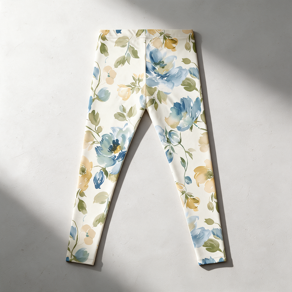 A pair of women's leggings laid flat on a light gray surface, featuring a watercolor-style floral print with blue, yellow, and green flowers and leaves on an off-white background.