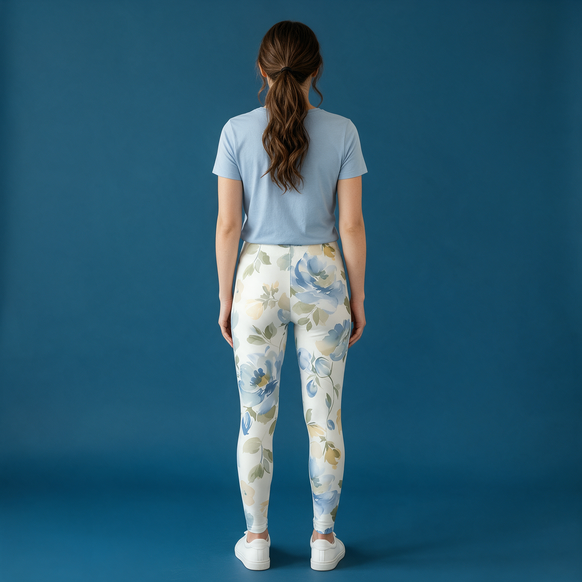 A woman stands with her back to the camera wearing floral print leggings with a light blue t-shirt, set against a solid blue studio background.