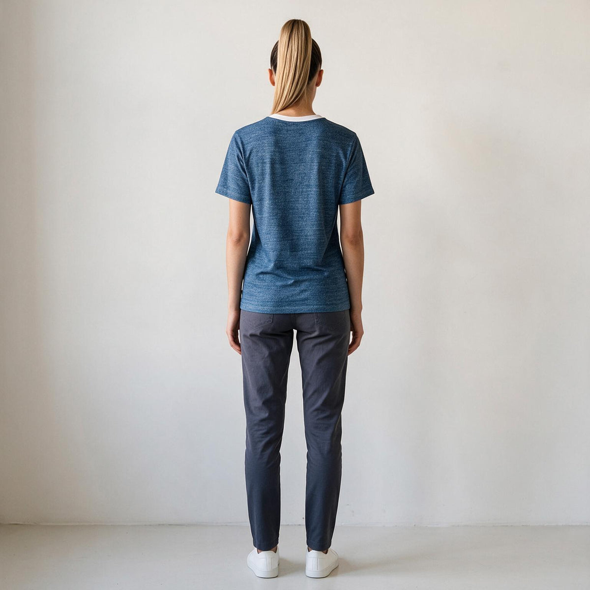 A woman is shown from behind wearing a heathered blue short-sleeve t-shirt, dark gray pants, and white sneakers, standing against a plain off-white wall.