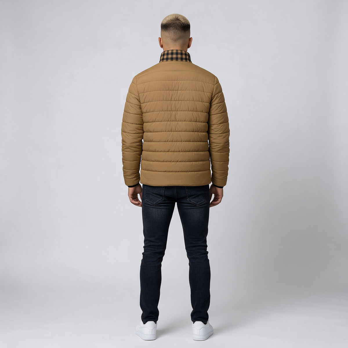 A man viewed from behind wearing a tan quilted puffer jacket with a black and beige plaid collar, dark blue jeans, and white sneakers, standing against a light gray studio background.
