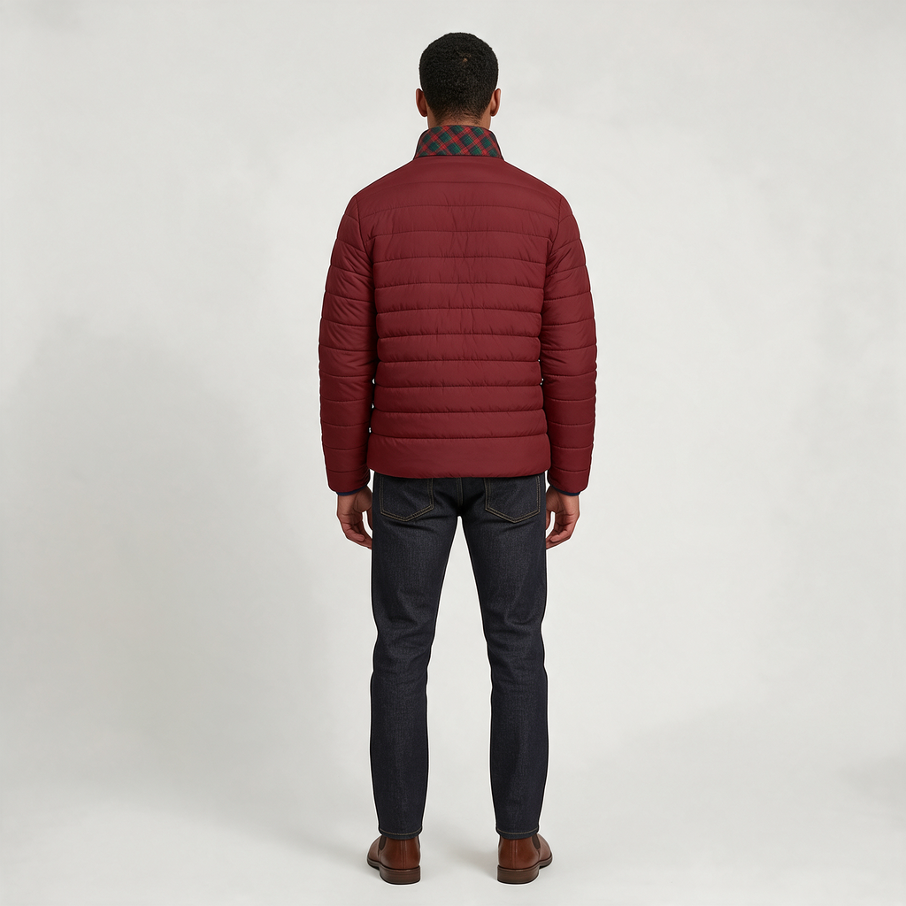 A man viewed from the back wearing a burgundy quilted puffer jacket over a dark plaid shirt, dark blue jeans, and brown leather shoes, set against a plain light gray studio background.
