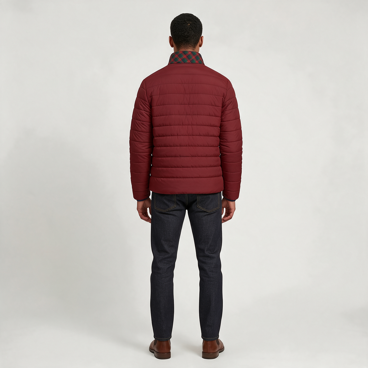 A man viewed from the back wearing a burgundy quilted puffer jacket over a dark plaid shirt, dark blue jeans, and brown leather shoes, set against a plain light gray studio background.