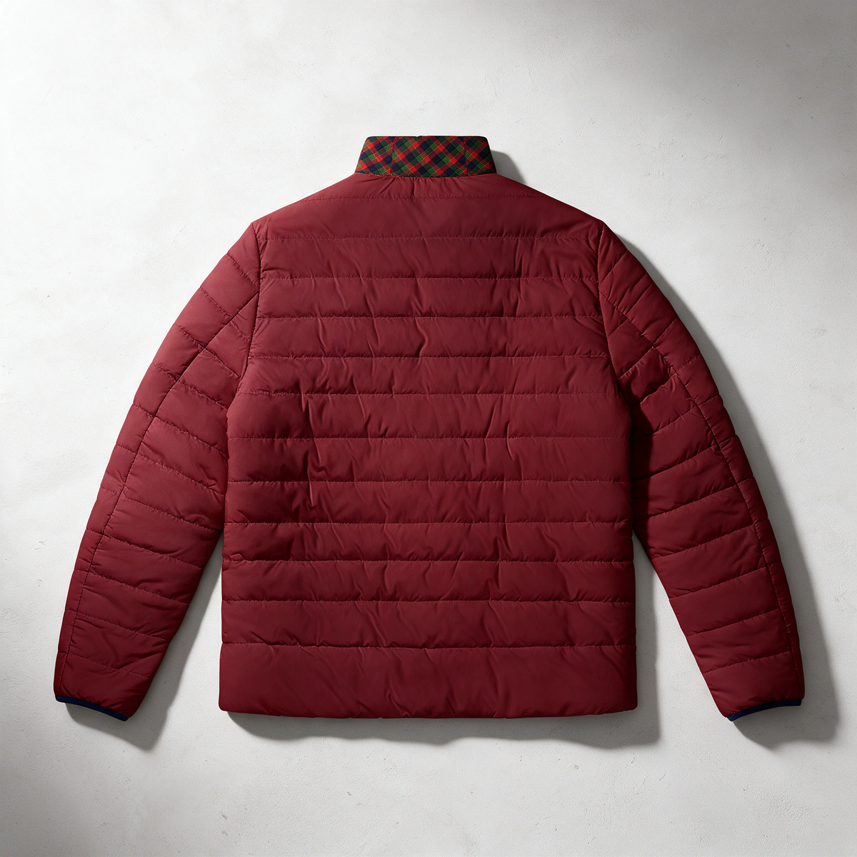 A burgundy quilted puffer jacket shown from the back, laid flat against a light gray textured background. The jacket features horizontal quilting, long sleeves, and a plaid patterned collar in red and green.