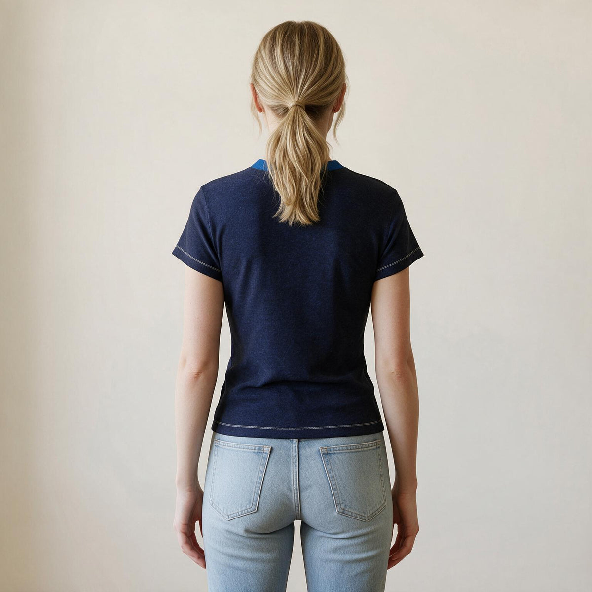A woman viewed from behind wearing a navy blue short-sleeve t-shirt with white contrast stitching and light-wash blue jeans, standing against a plain beige wall.