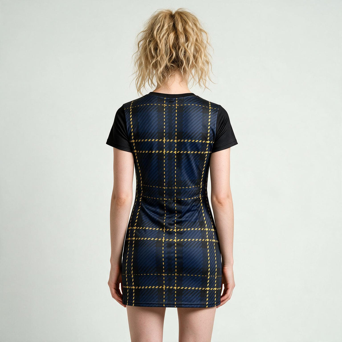 A woman viewed from the back wearing a short-sleeve athletic mini dress with a navy blue grid pattern and yellow dashed lines, set against a plain light gray background.