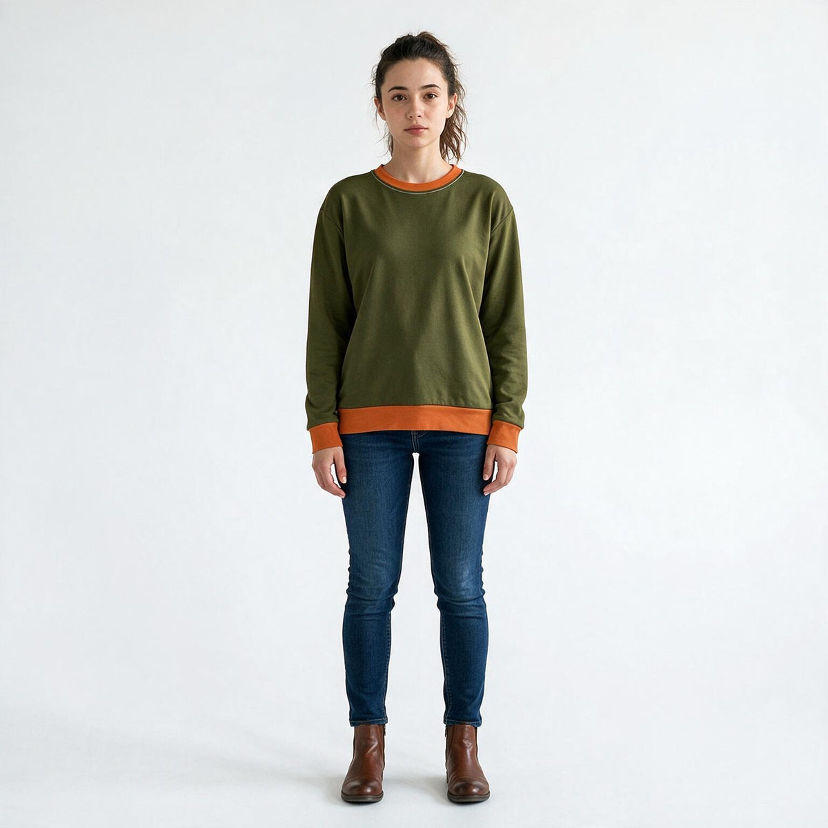 A woman stands against a plain white studio background wearing an olive green crewneck sweatshirt with contrasting orange ribbed trim at the collar, cuffs, and hem, paired with dark blue skinny jeans and brown ankle boots.
