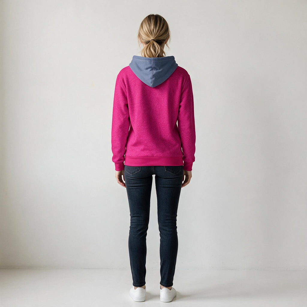 A woman viewed from behind wearing a bright pink hoodie with a contrasting blue hood, paired with dark blue jeans and white sneakers, standing against a plain light gray studio background.