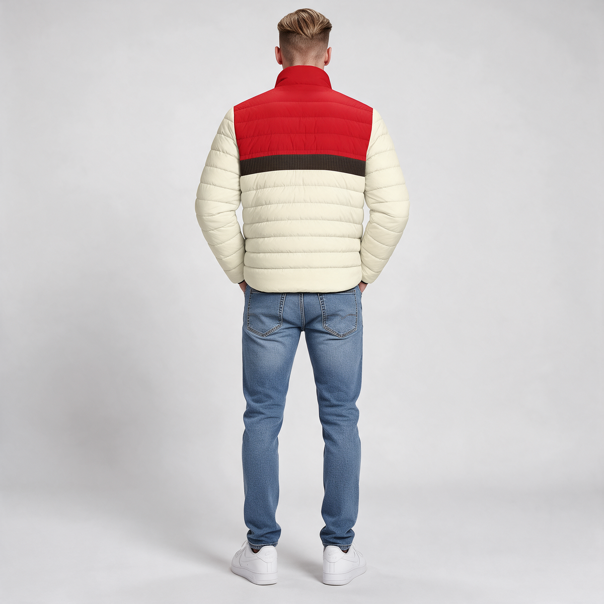 A man viewed from behind wearing a color-blocked puffer jacket with cream, red, and dark brown horizontal stripes, paired with blue jeans and white sneakers, against a light gray studio background.