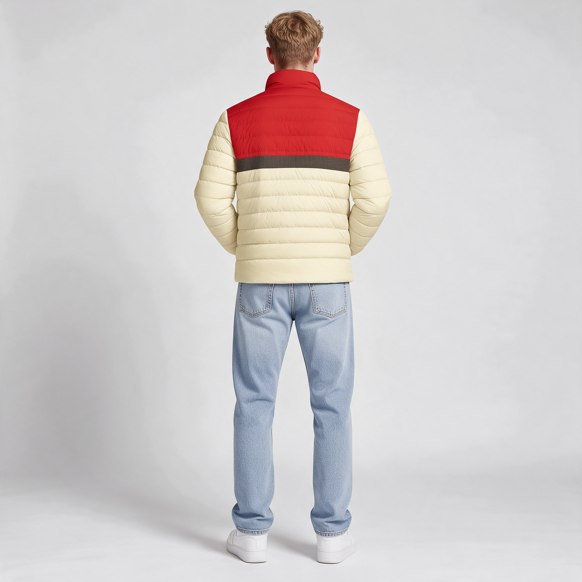A man viewed from behind wearing a color-blocked puffer jacket with a cream body, a red upper back and collar, and a dark gray horizontal stripe, paired with light blue jeans and white sneakers on a light gray studio background.