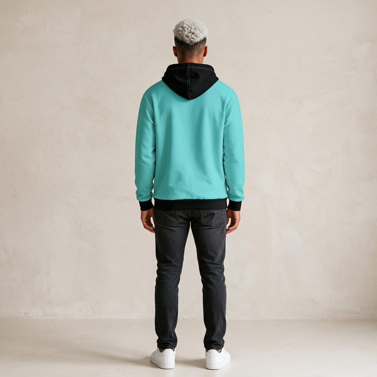 A person viewed from the back wearing a turquoise hoodie with a black hood and black ribbed trim, paired with dark jeans and white sneakers, standing against a plain beige wall.