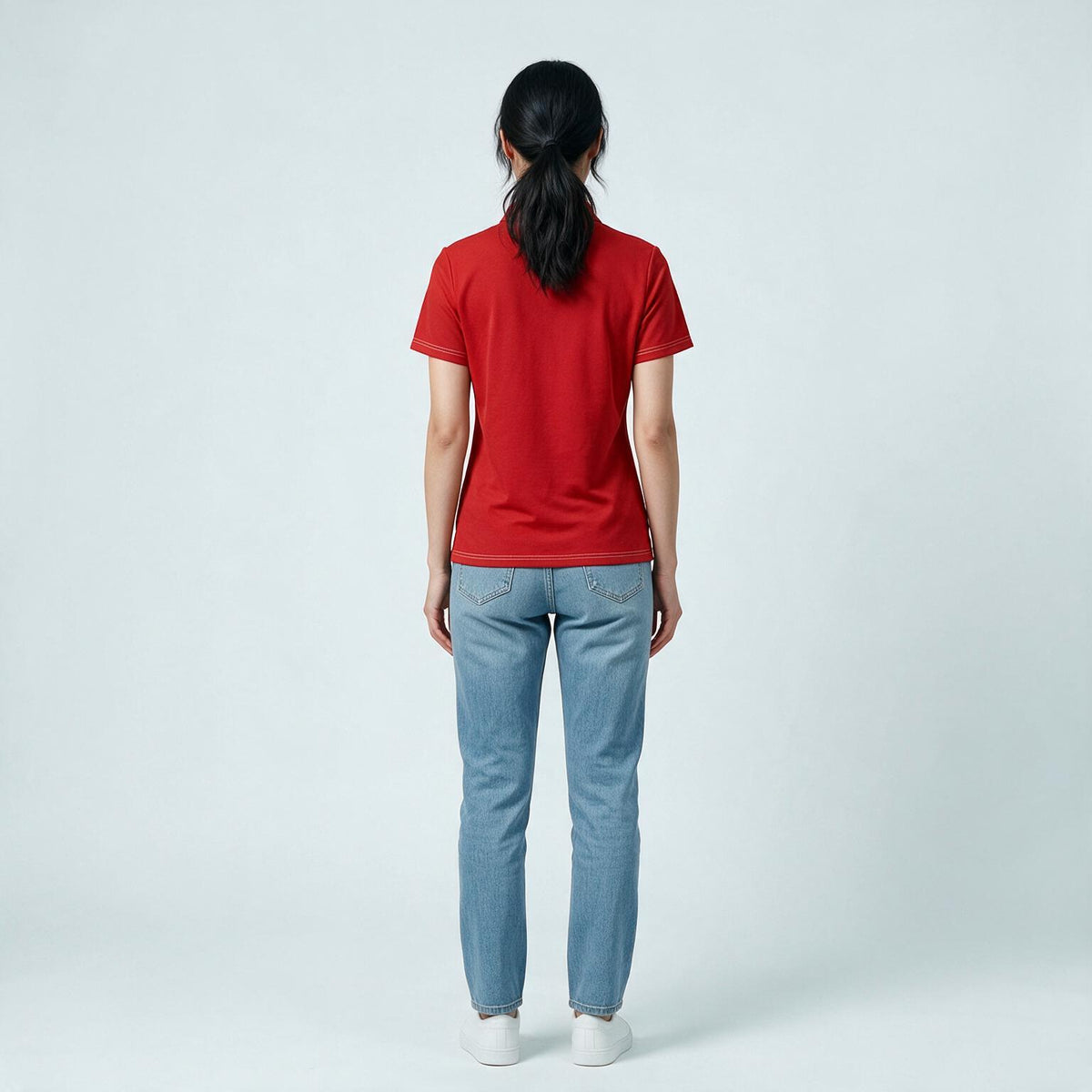 A woman is shown from behind wearing a red short-sleeve t-shirt, light blue jeans, and white sneakers against a light gray studio background.