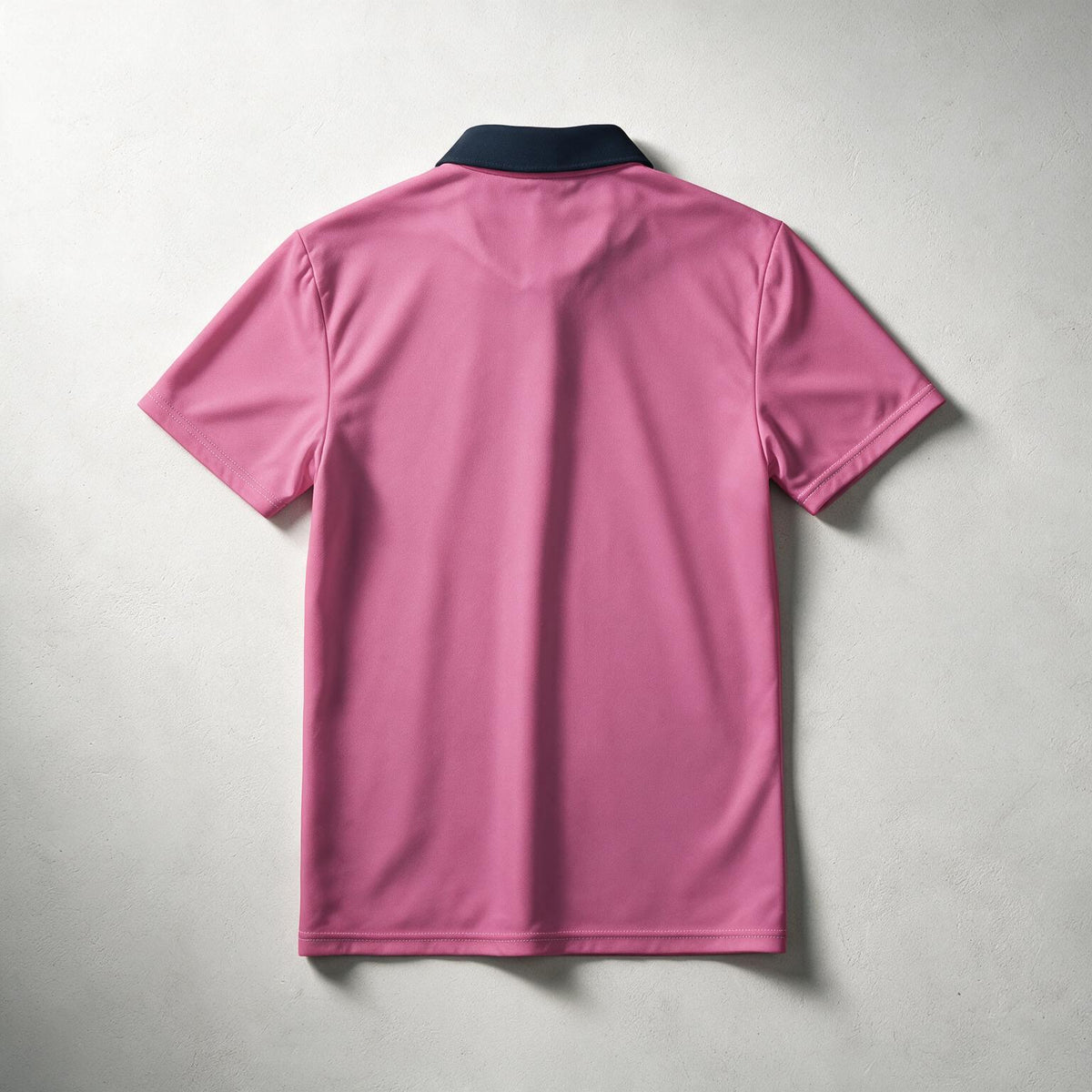A pink short-sleeve polo shirt shown from the back, featuring a contrasting dark navy collar, laid flat against a textured light gray background.