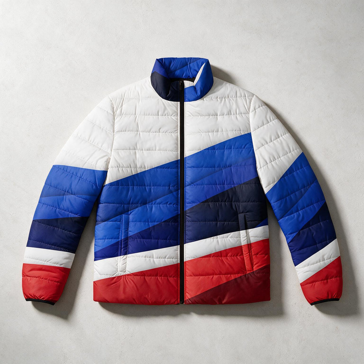 A quilted puffer jacket with a geometric color-blocked pattern in white, blue, navy, and red, laid flat against a light gray textured background.