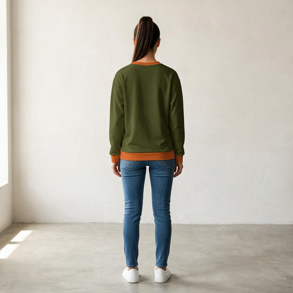 A woman viewed from behind wearing an olive green crewneck sweatshirt with orange ribbed trim, blue skinny jeans, and white sneakers, standing in a minimalist room with a white wall and concrete floor.