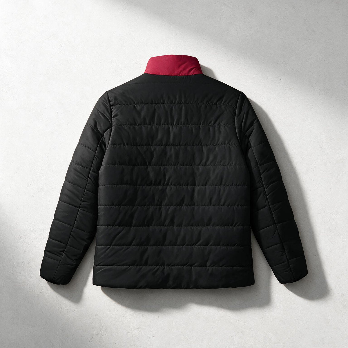 A black quilted puffer jacket shown from the back, featuring a contrasting red collar, photographed against a light gray textured background with soft shadows.