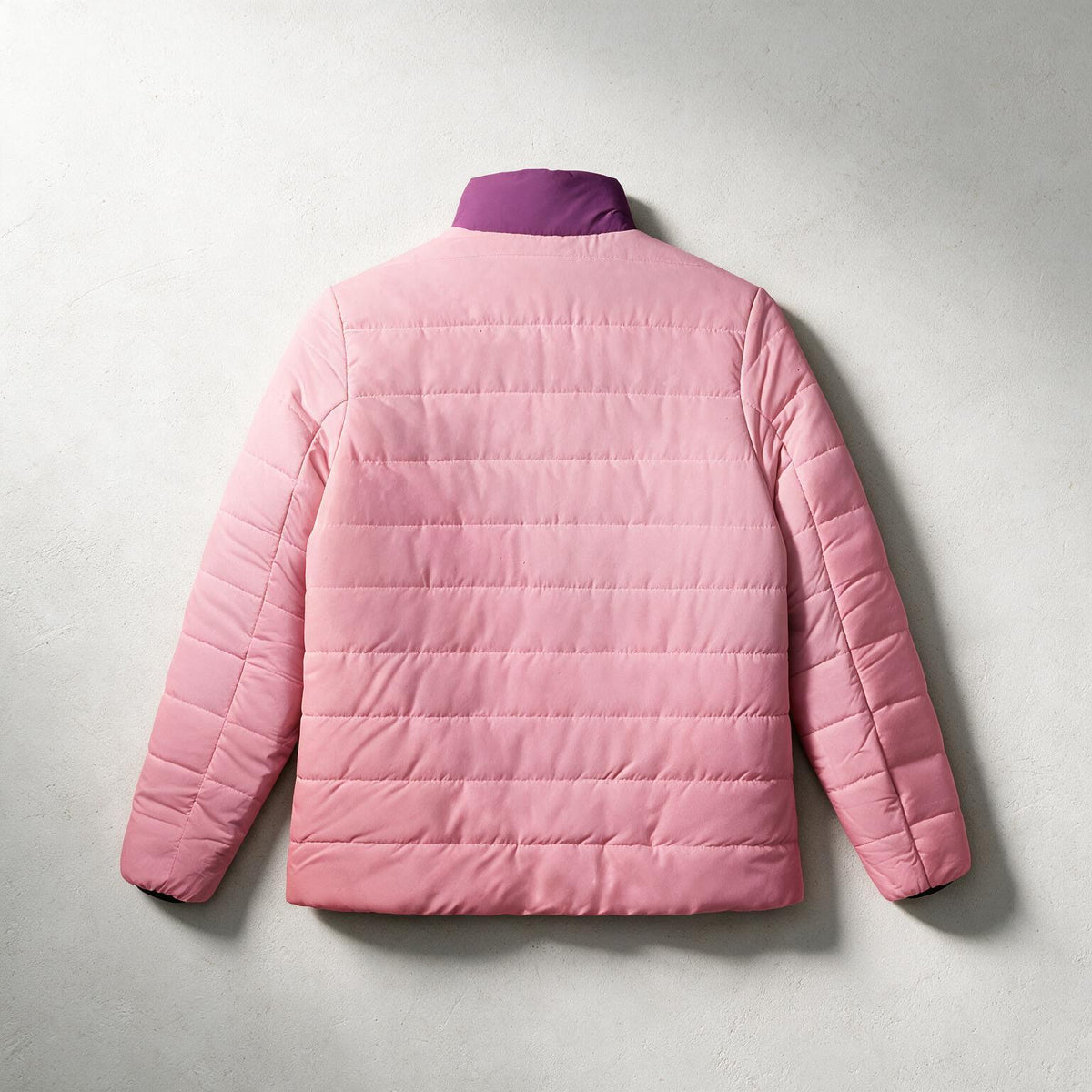 A light pink quilted puffer jacket shown from the back, featuring a contrasting dark purple collar, laid flat against a textured light gray background.