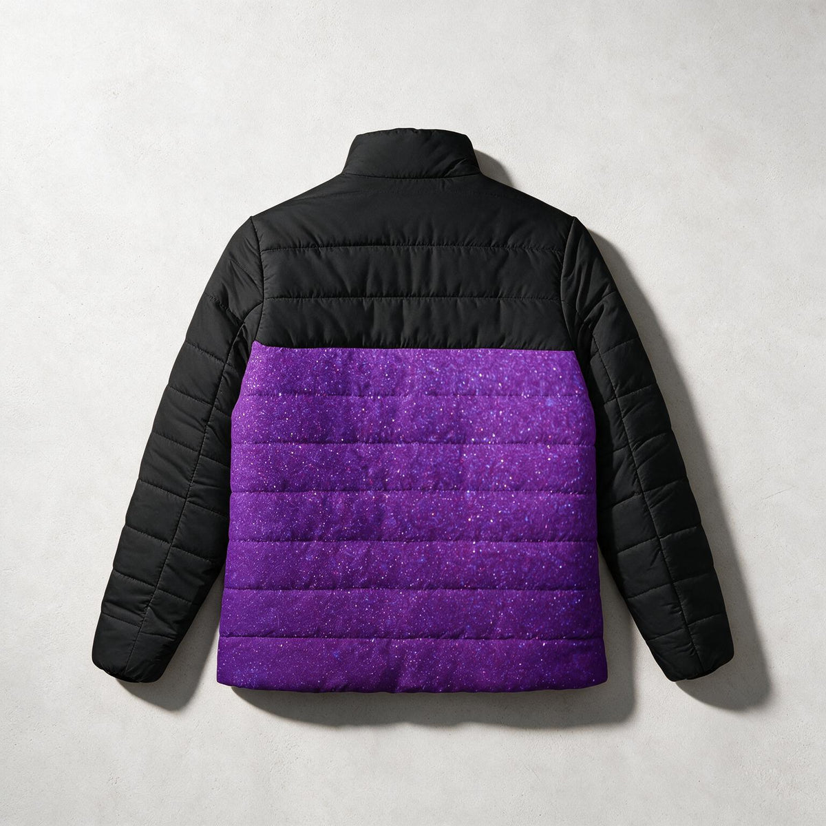 A children's puffer jacket shown from the back, featuring a solid black quilted upper section and sleeves, and a vibrant purple glitter-textured lower section, laid flat on a light gray background.
