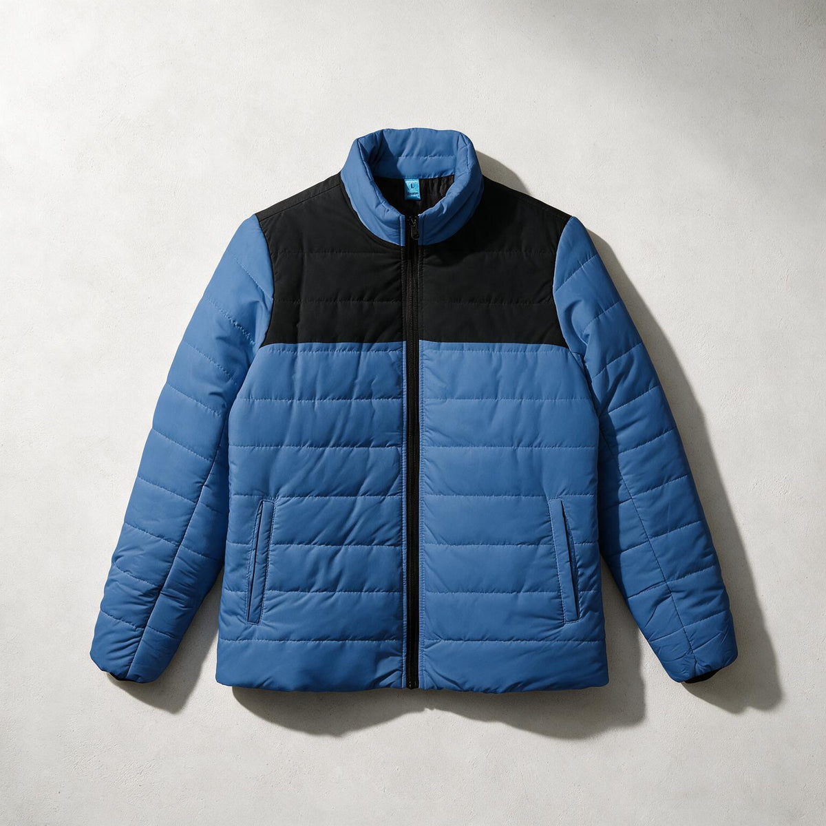 A color-blocked puffer jacket with a blue body and sleeves, and a black yoke across the chest, laid flat on a light gray textured background.