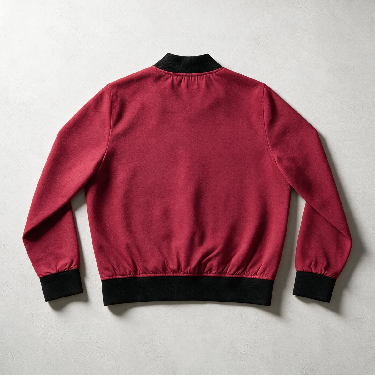 A burgundy bomber jacket shown from the back, featuring black ribbed collar, cuffs, and hem, laid flat on a light gray textured background.