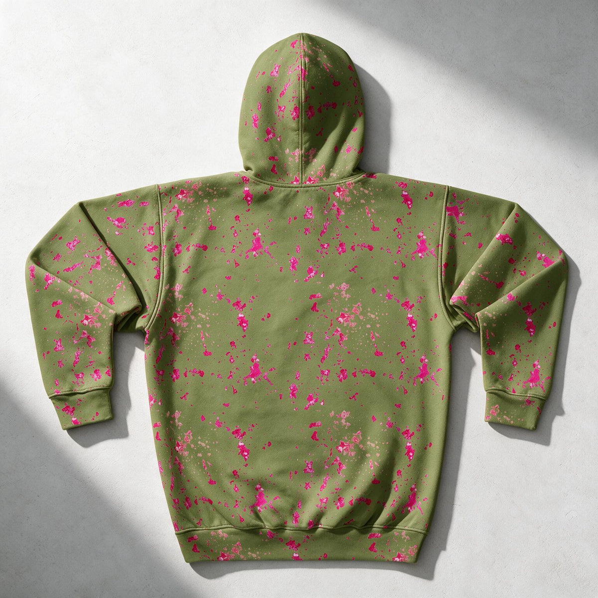 An olive green hoodie with a vibrant pink splatter print, shown from the back on a light gray textured background with soft diagonal shadows.