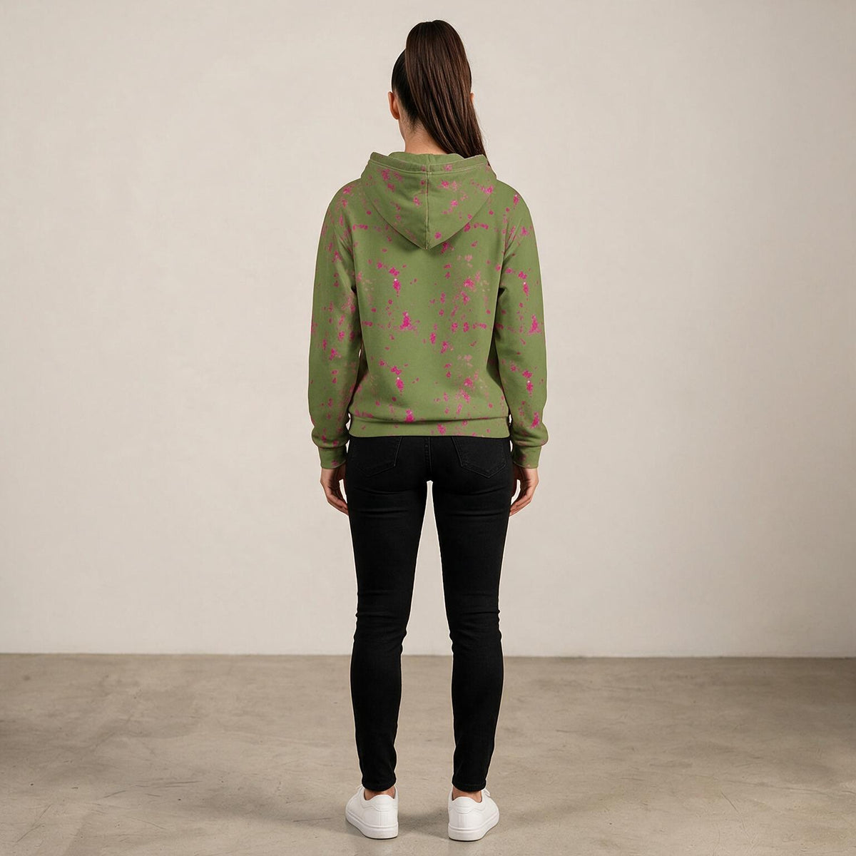 A woman viewed from behind wearing an olive green hoodie with a pink floral pattern, paired with black skinny jeans and white sneakers, standing against a neutral beige wall.