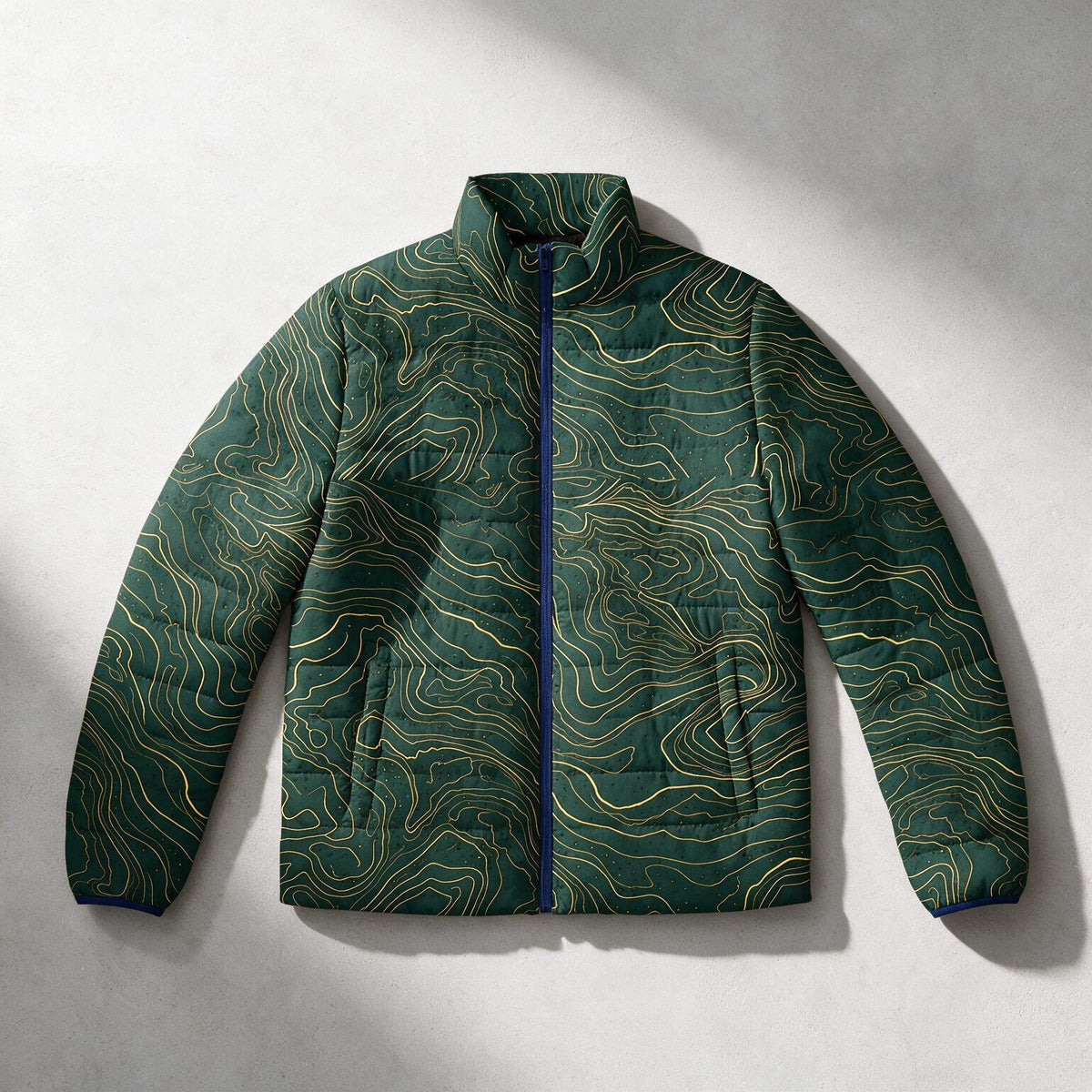 A dark green quilted puffer jacket with a gold topographic contour line pattern, laid flat on a light gray textured background with soft shadows.