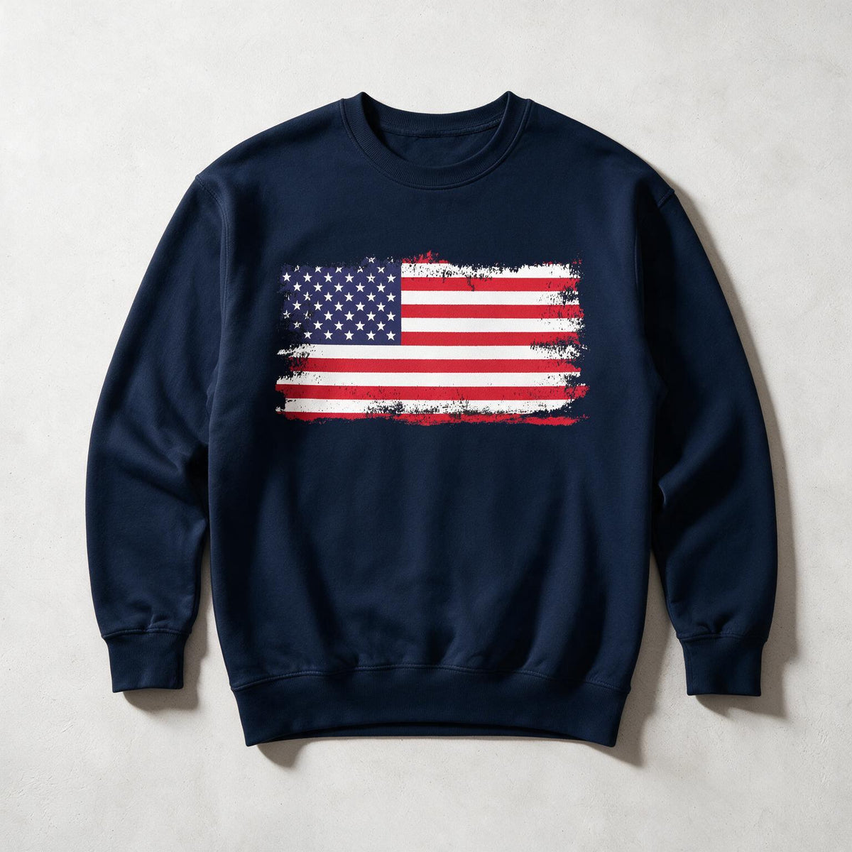 A navy blue crewneck sweatshirt featuring a distressed American flag graphic printed on the chest, laid flat against a light gray textured background.