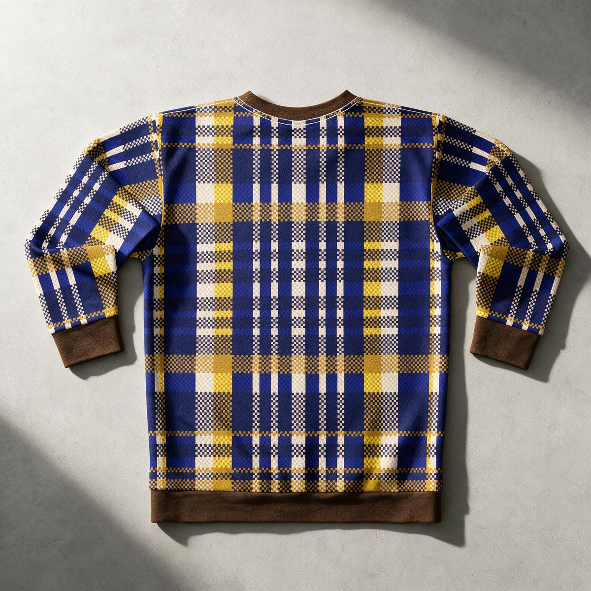 A long-sleeve crewneck sweatshirt with a blue, yellow, and white plaid pattern, featuring solid brown ribbed cuffs, hem, and collar, laid flat on a textured gray background.