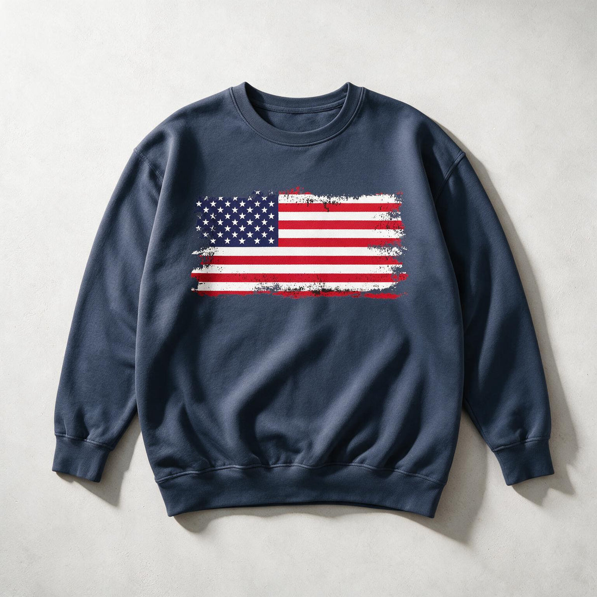 A navy blue crewneck sweatshirt featuring a distressed American flag graphic on the chest, laid flat on a light gray textured background.