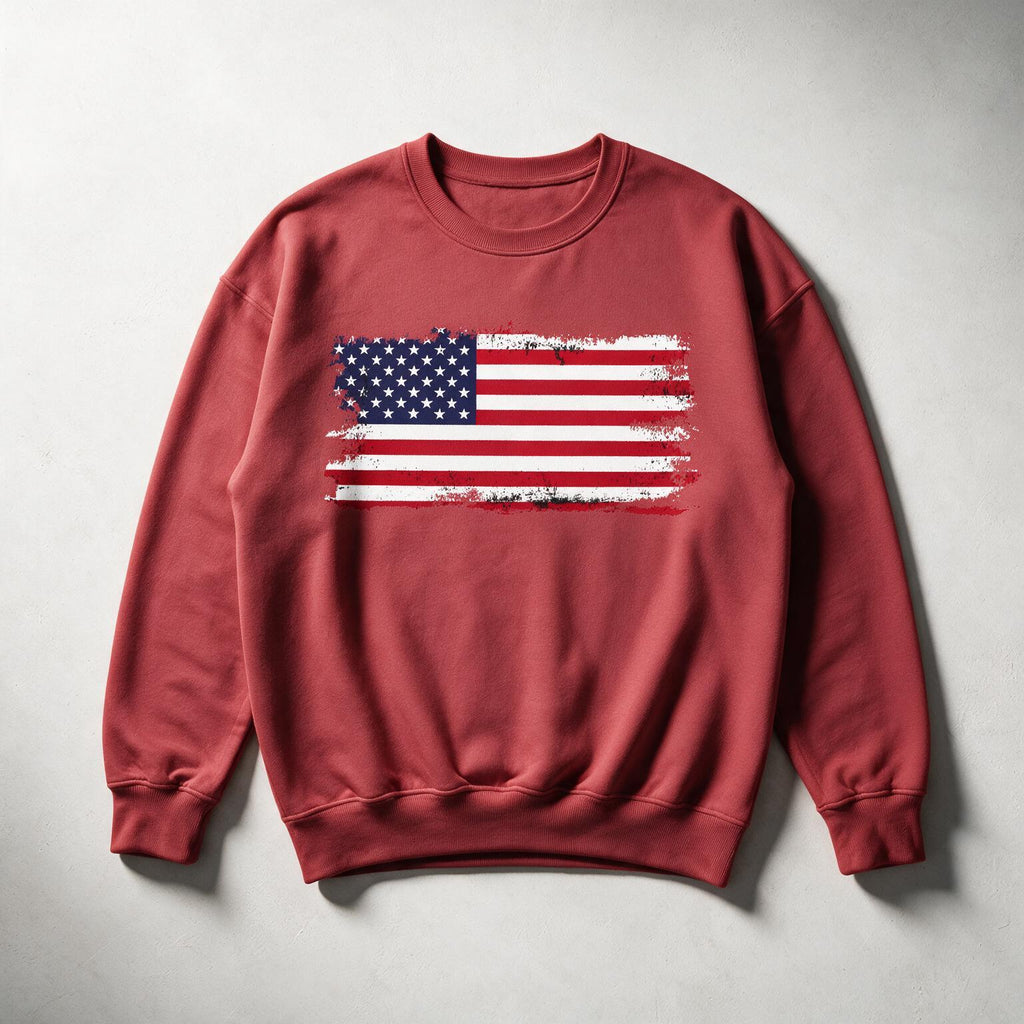 A crewneck sweatshirt in a muted brick red color, featuring a distressed American flag graphic printed across the chest. The sweatshirt is laid flat on a neutral gray background.