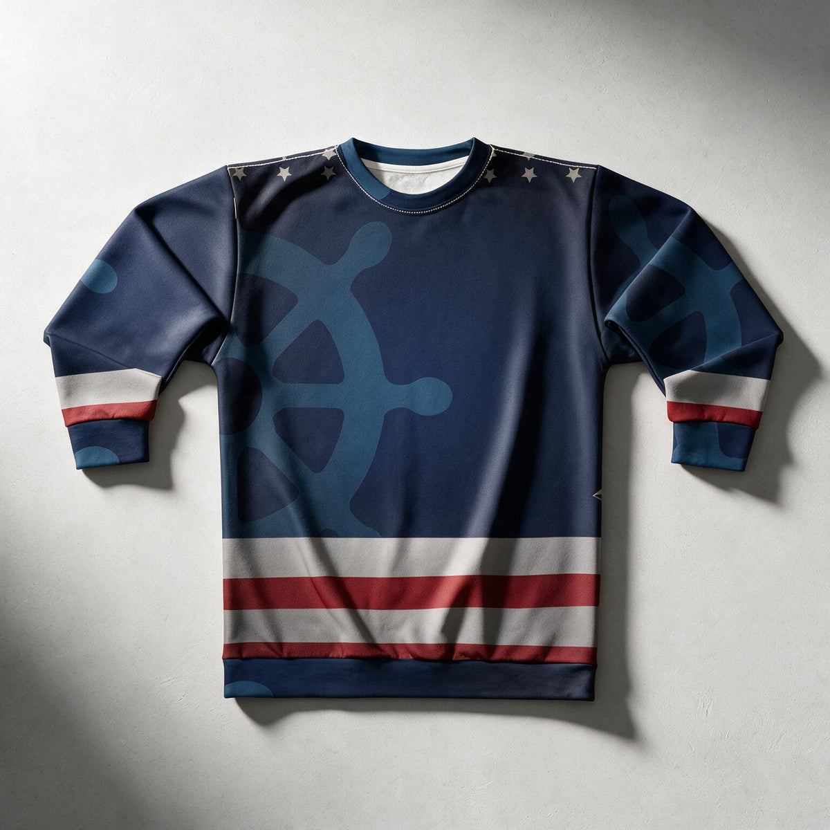 A crewneck sweatshirt featuring a navy blue and light blue ship's wheel graphic, with red and white horizontal stripes near the hem and cuffs, and small white stars on the shoulders, laid flat on a gray background.
