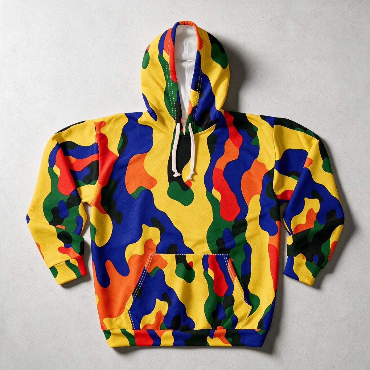 A long-sleeve pullover hoodie with a bold, multicolor abstract camouflage pattern featuring yellow, blue, red, green, and orange shapes, laid flat on a light gray textured background.