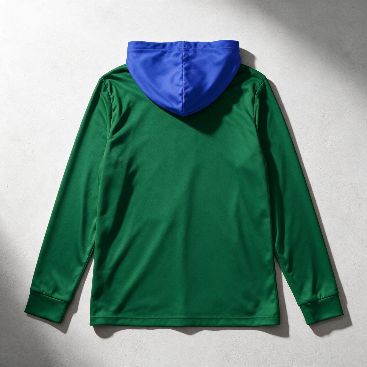 A green hooded sweatshirt shown from the back, featuring a contrasting royal blue hood, laid flat on a light gray textured background with dramatic diagonal shadows.