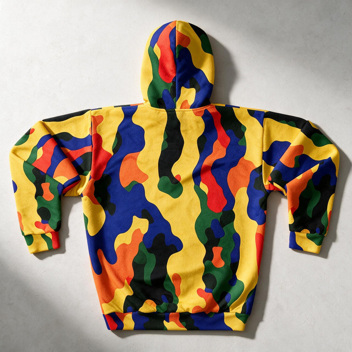 A hooded sweatshirt shown from the back, featuring a bold multicolor camouflage pattern with yellow, blue, green, orange, and black shapes, laid flat on a light gray background.