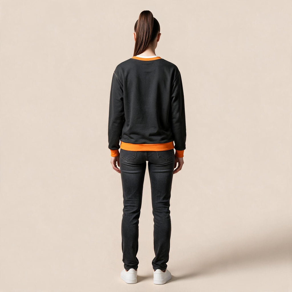 A woman viewed from the back wearing a black crewneck sweatshirt with bright orange ribbed trim at the collar, cuffs, and hem, paired with dark gray jeans and white sneakers, against a solid beige studio background.