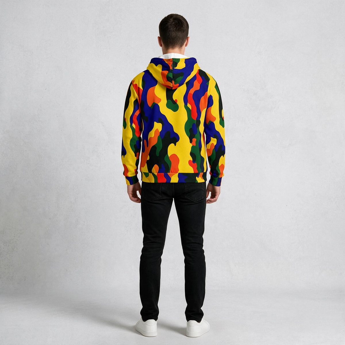 A man viewed from behind wearing a vibrant multicolor camouflage hoodie with yellow, blue, red, green, and orange shapes, paired with black pants and white sneakers, against a light gray textured studio background.