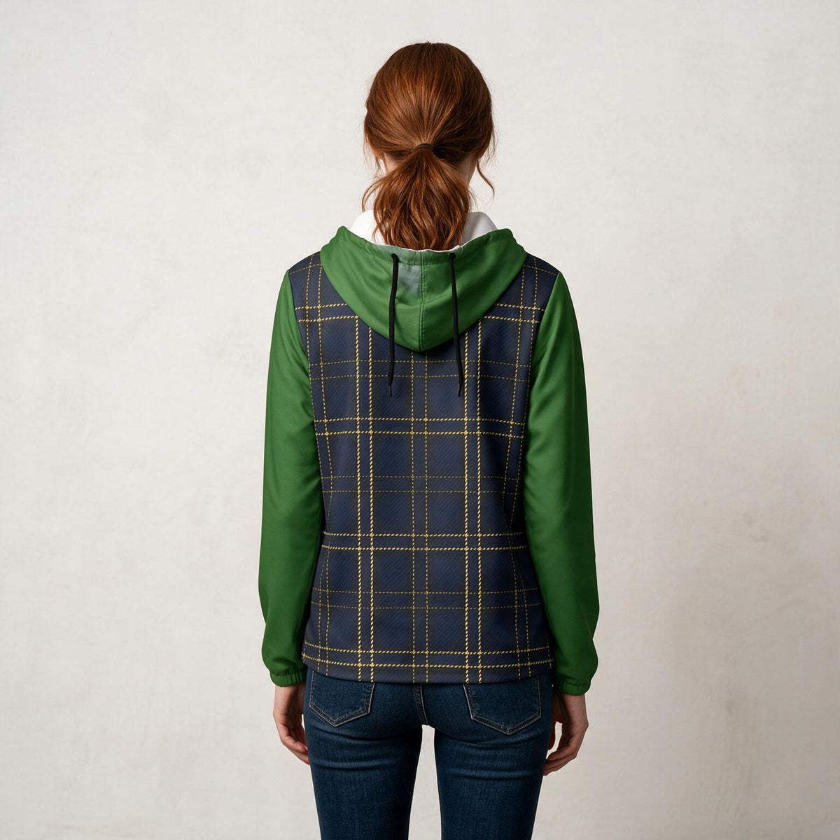 A woman viewed from behind wearing a hooded pullover with green sleeves and a navy plaid body with thin gold lines, paired with dark blue jeans against a light textured wall.